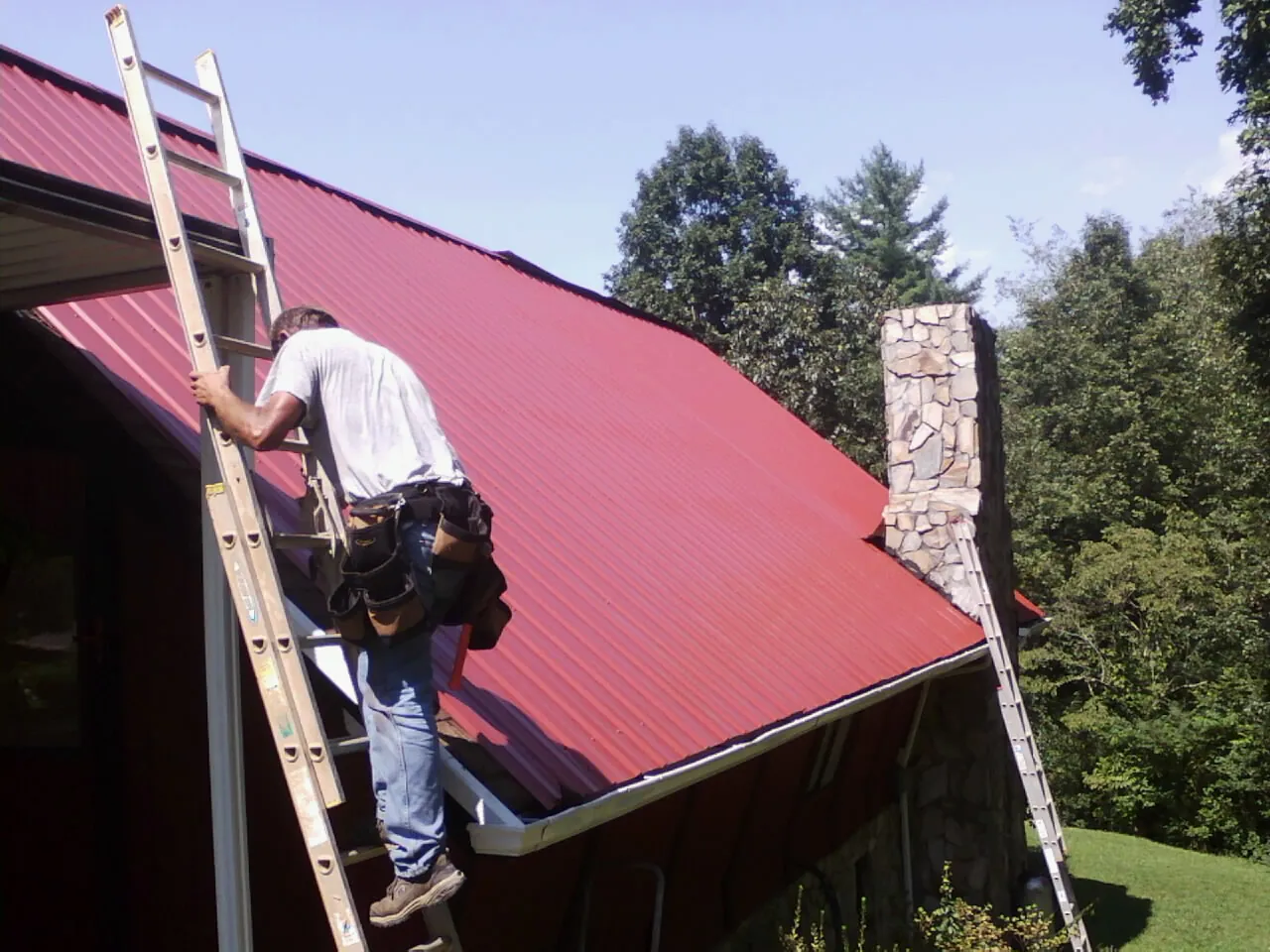 Roofer on a ladder beside a red metal roof and stone chimney — Soffit & Fascia Repair by Roofing Brothers in Brookings